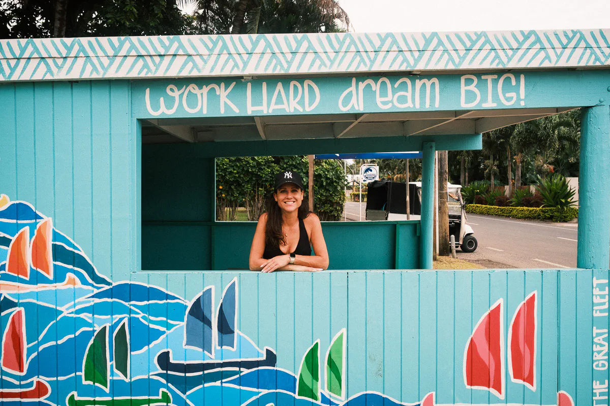 Sam leaning out the window of a colourful blue food stall painted with sailboats and the words "Work Hard Dream Big", Rarotonga